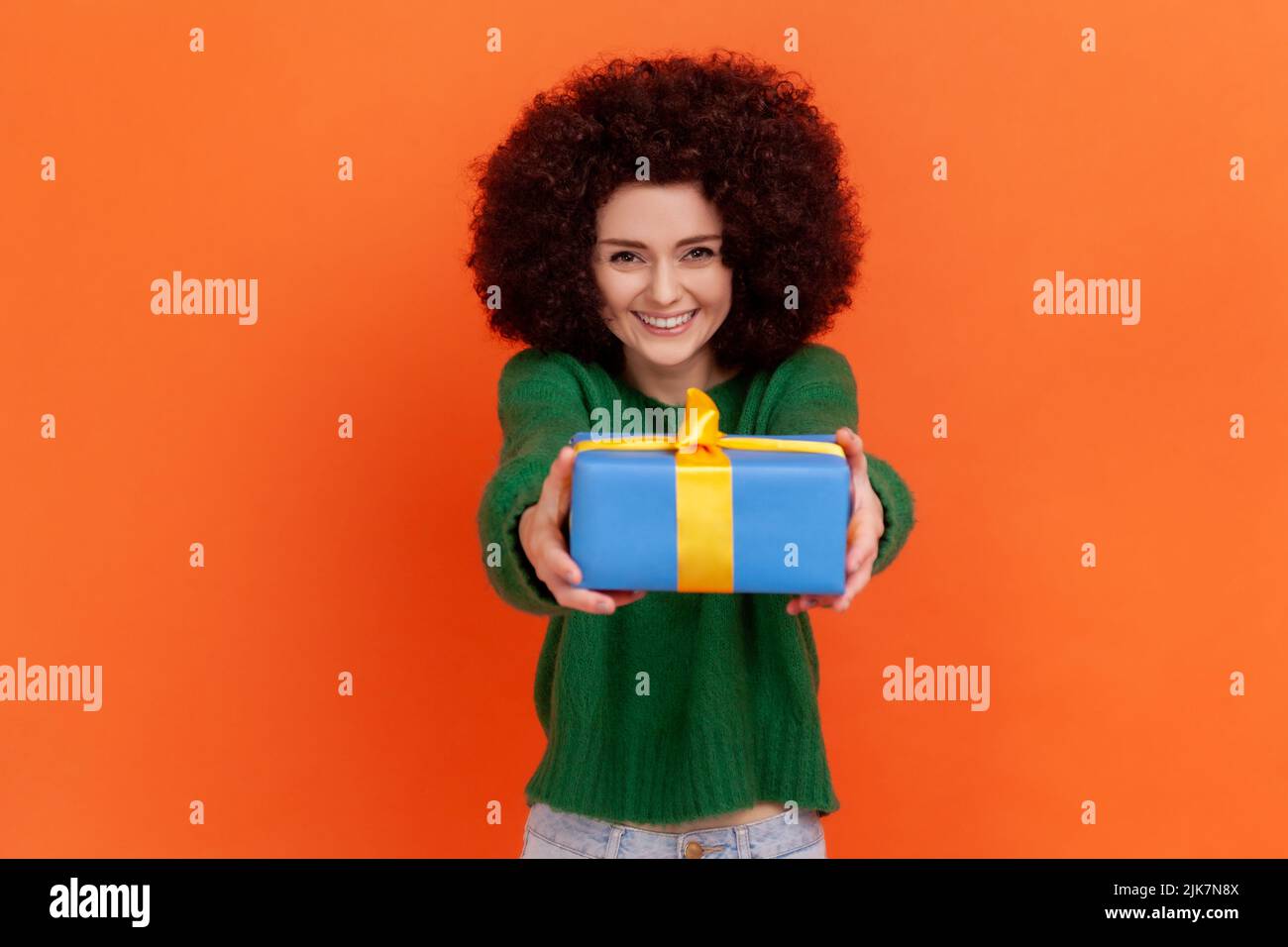 Portrait of winsome woman with Afro hairstyle wearing green casual style sweater giving present, holding out blue wrapped gift box. Indoor studio shot isolated on orange background. Stock Photo