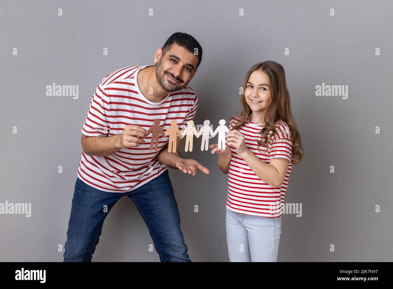 Portrait of father and daughter in striped T-shirts holding paper chain multiracial people, symbol of community, unity, people and support. Indoor studio shot isolated on gray background. Stock Photo