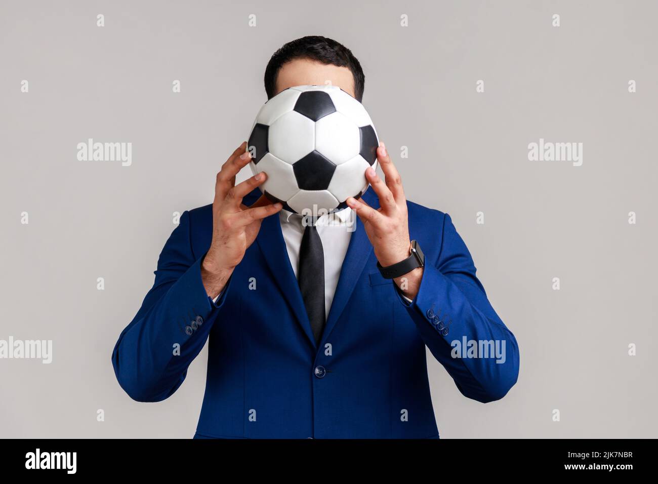 Portrait of unknown anonymous businessman supporting his favorite team covering face with soccer ball, wearing official style suit. Indoor studio shot isolated on gray background. Stock Photo