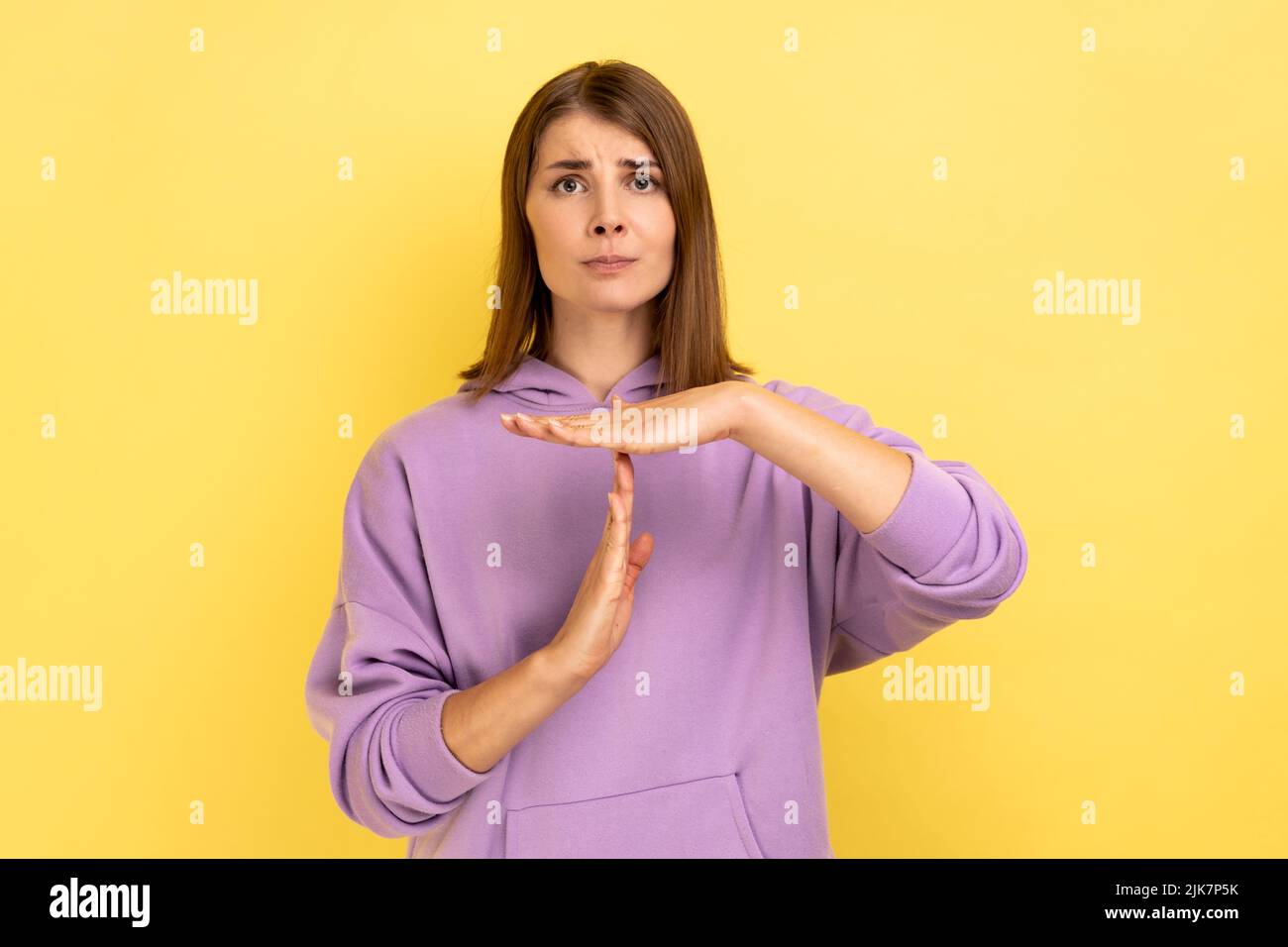 I need more time. Portrait of displeased woman showing time out hand gesture, looking imploringly, worried about deadline, wearing purple hoodie. Indoor studio shot isolated on yellow background. Stock Photo