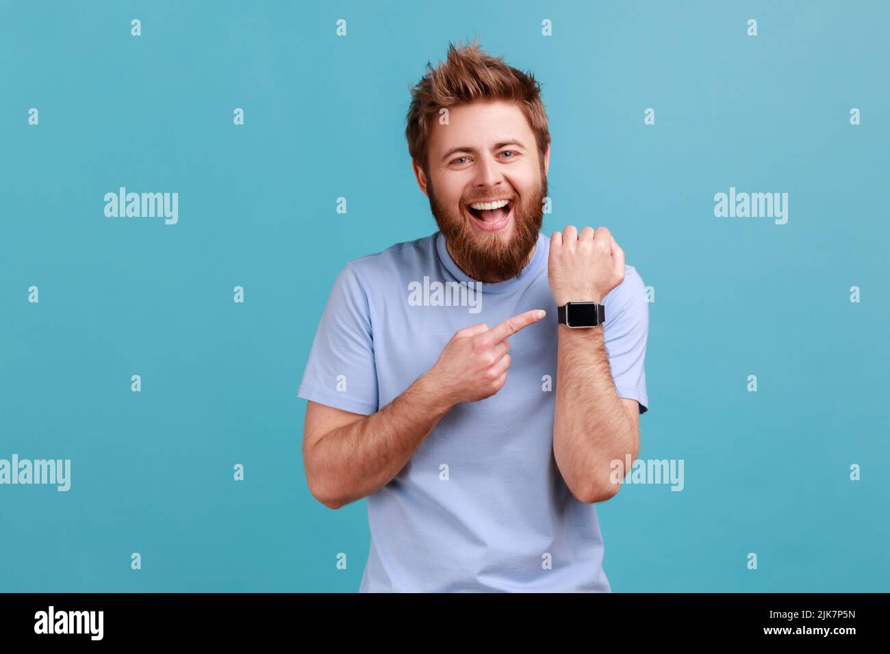 Portrait of positive funny bearded man standing with open mouth and satisfied expression, pointing with finger ar smartwatch and laughing. Indoor studio shot isolated on blue background. Stock Photo