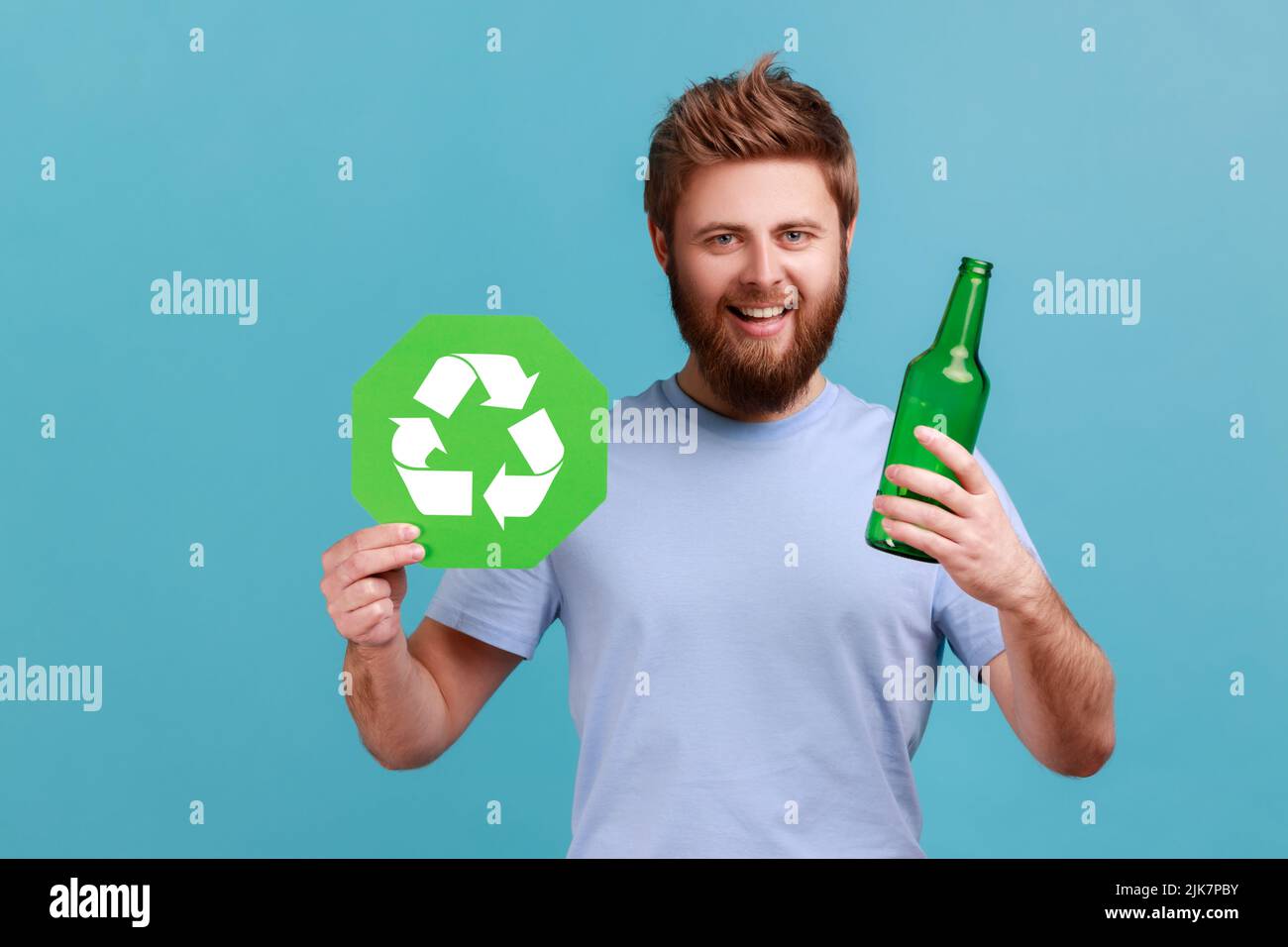 Portrait of smiling positive attractive bearded man holding glass bottle and recycling green symbol, sorting his rubbish, saving ecology. Indoor studio shot isolated on blue background. Stock Photo