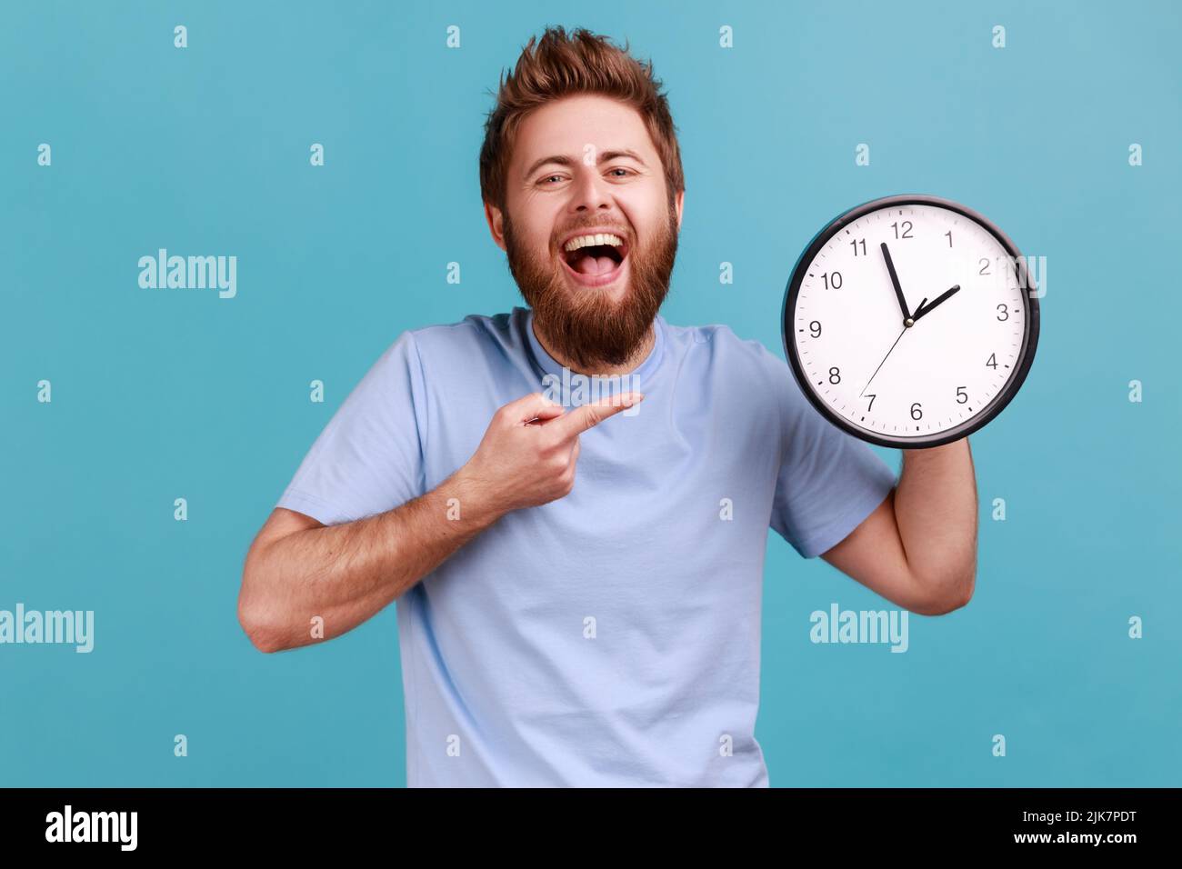 Portrait of happy positive handsome bearded man pointing at wall clock with finger, looking at camera with toothy smile, laughing. Indoor studio shot isolated on blue background. Stock Photo