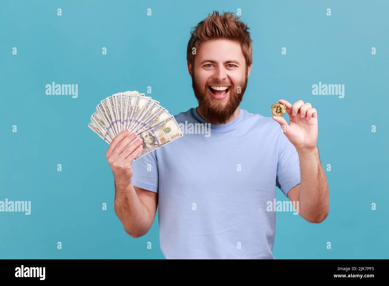 Portrait of positive optimnistic handsome bearded man showing dollar banknotes and bitcoin, digital money, electronic commerce, expressing excitement. Indoor studio shot isolated on blue background. Stock Photo