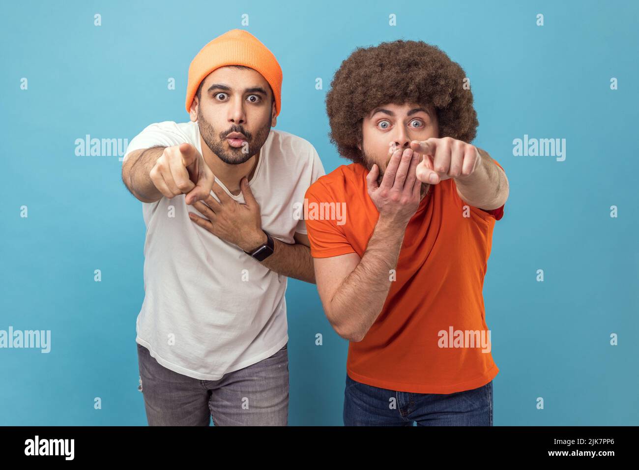 Wow, look at this. Portrait of two astonished surprised young adult hipster men looking at camera with big eyes, pointing at camera with fingers. Indoor studio shot isolated on blue background. Stock Photo