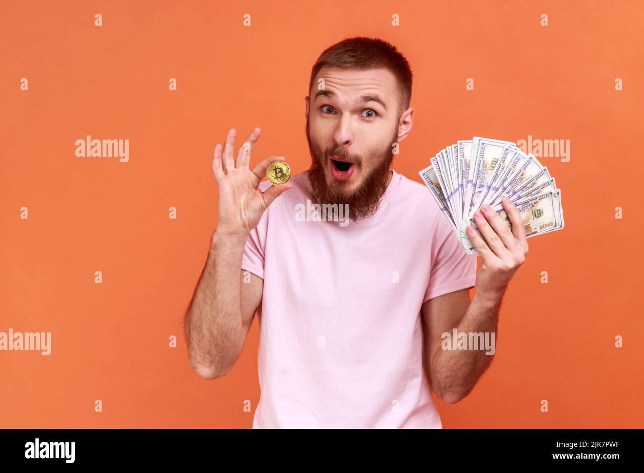 Portrait of bearded man holding bitcoin and dollar banknotes, looking at camera, astonished profit from cryptocurrency, wearing pink T-shirt. Indoor studio shot isolated on orange background. Stock Photo