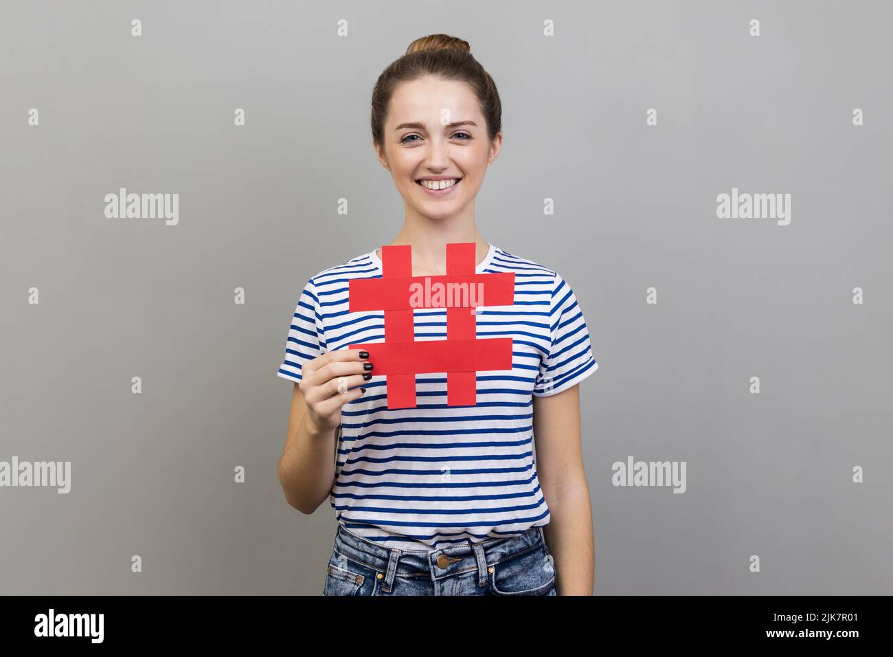 Hashtag and blogging. Portrait of smiling positive optimistic woman wearing striped T-shirt holding big paper hash symbol and smiling at camera. Indoor studio shot isolated on gray background. Stock Photo