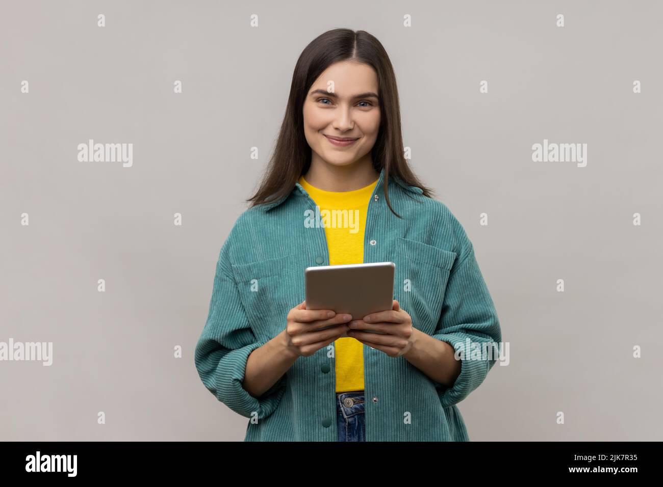 Woman with dark hair working online, using tablet for checking social networks, looking at camera with positive expression, wearing casual style jacket. Indoor studio shot isolated on gray background. Stock Photo