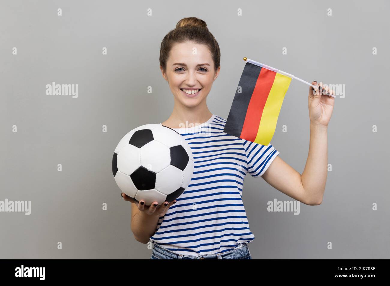 Portrait of joyful cheerful woman wearing striped T-shirt holding soccer ball and german flag, looking smiling at camera, cheering up. Indoor studio shot isolated on gray background. Stock Photo
