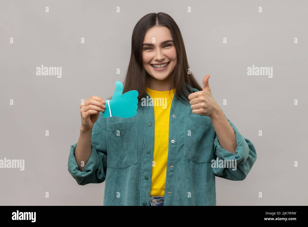 Woman blogger holding paper blue thumbs up sign, asking to rate her posts in social networks, happy satisfied expression, wearing casual style jacket. Indoor studio shot isolated on gray background. Stock Photo