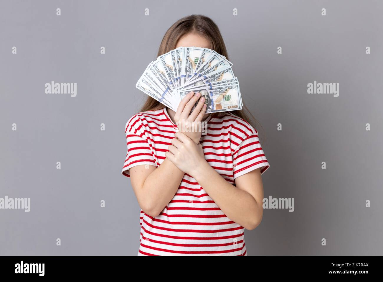Portrait of unknown anonymous rich little girl wearing striped T-shirt hiding her face behind dollars banknotes, income of money. Indoor studio shot isolated on gray background. Stock Photo