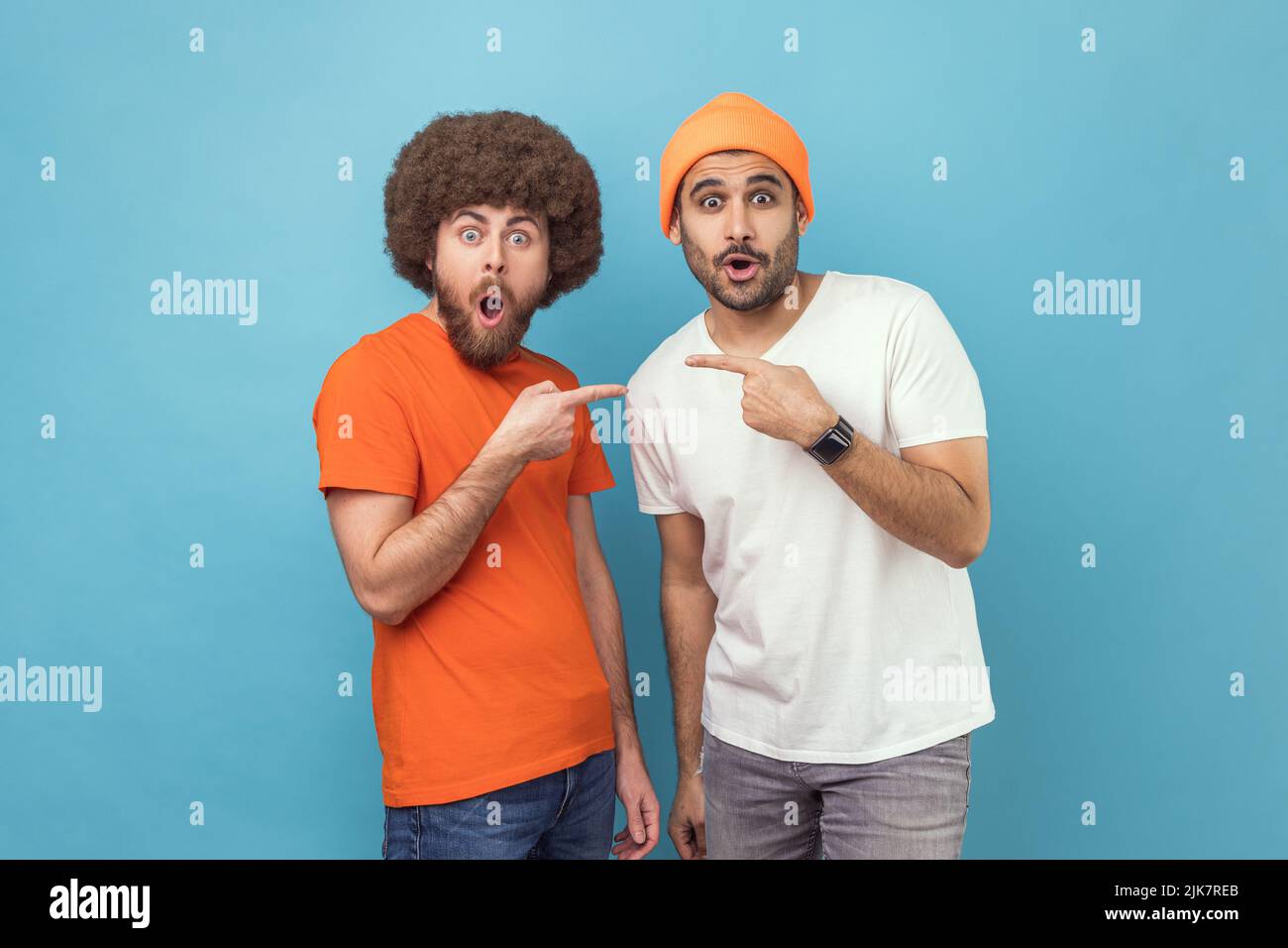 Portrait of two young adult hipster men pointing finger each other and looking at camera with shocked facial expressions and open mouth. Indoor studio shot isolated on blue background. Stock Photo