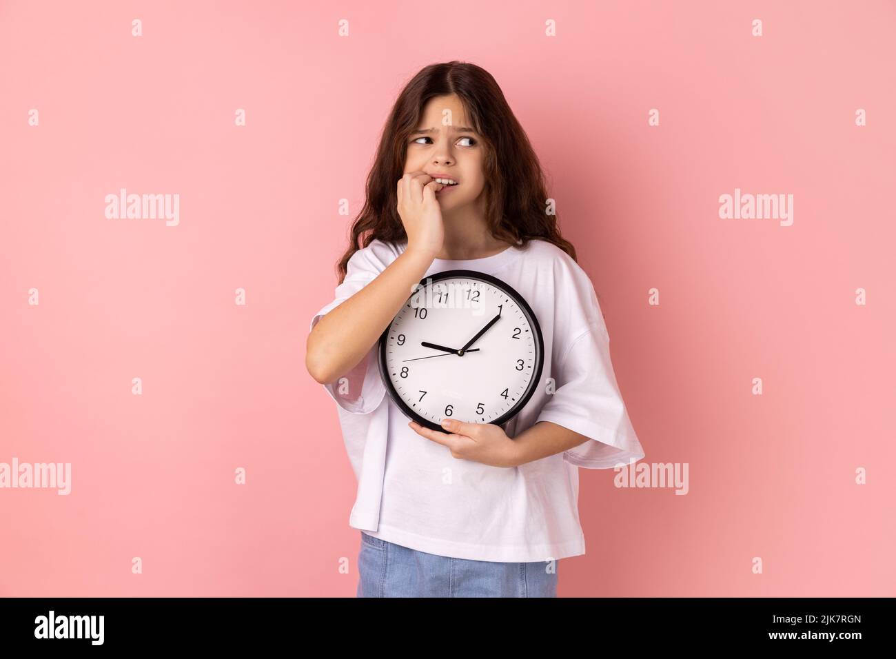 Portrait of nervous anxious little girl wearing white T-shirt biting nails on fingers holding big wall clock in hand, worried about deadline. Indoor studio shot isolated on pink background. Stock Photo