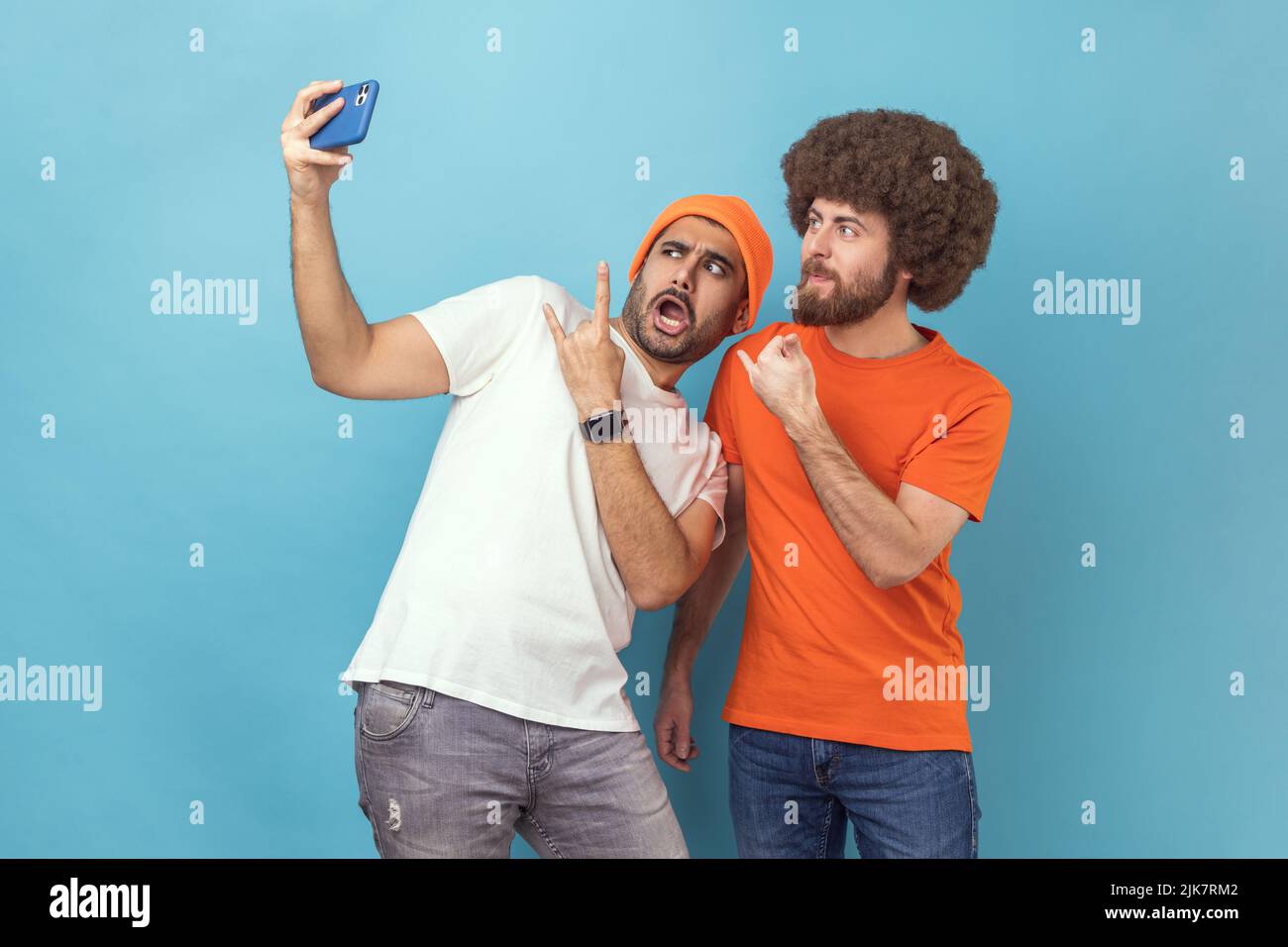 Portrait of funny cool two young adult hipster men blogger broadcasting livestream or taking selfie, showing rock and roll and shaka gestures. Indoor studio shot isolated on blue background. Stock Photo