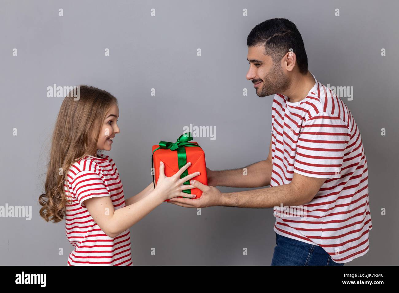 Portrait of satisfied father and daughter in striped T-shirts, man giving present box to his cute child, celebrating birthday together. Indoor studio shot isolated on gray background. Stock Photo