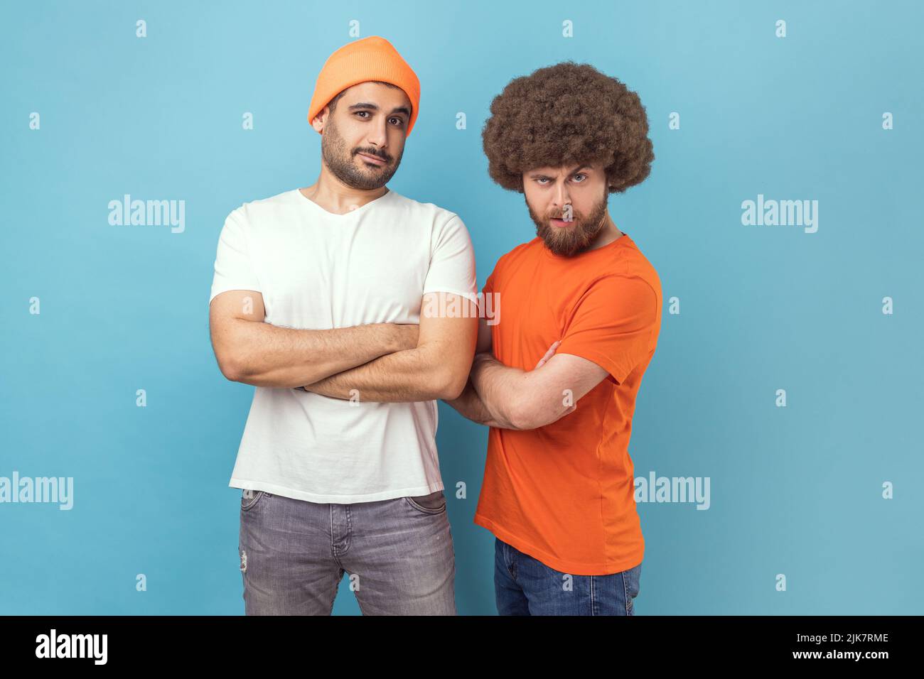 Portrait of two handsome serious young adult hipster men standing looking at camera with assertive expression, keeping hands folded. Indoor studio shot isolated on blue background. Stock Photo