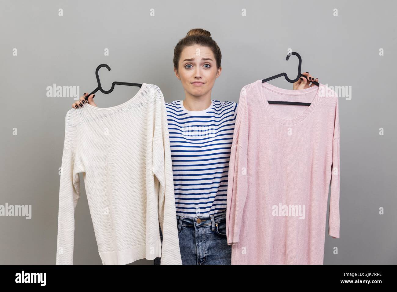 Portrait of confused puzzled woman wearing striped T-shirt holding two shirts on hangers, choosing best look, looking at camera with doubtful expression. Indoor studio shot isolated on gray background Stock Photo