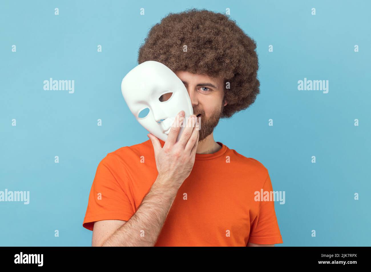 Portrait of man with Afro hairstyle wearing orange T-shirt peeping out white face mask, hiding his real feelings, pretending to be another person. Indoor studio shot isolated on blue background. Stock Photo