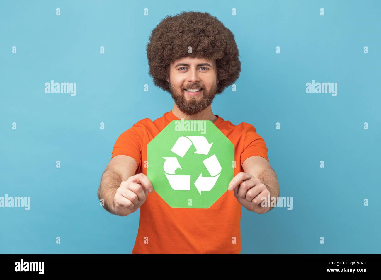 Portrait of man with Afro hairstyle in orange T-shirt holding waste recycling symbol in his hand, worrying about ecology and environmental pollution. Indoor studio shot isolated on blue background. Stock Photo