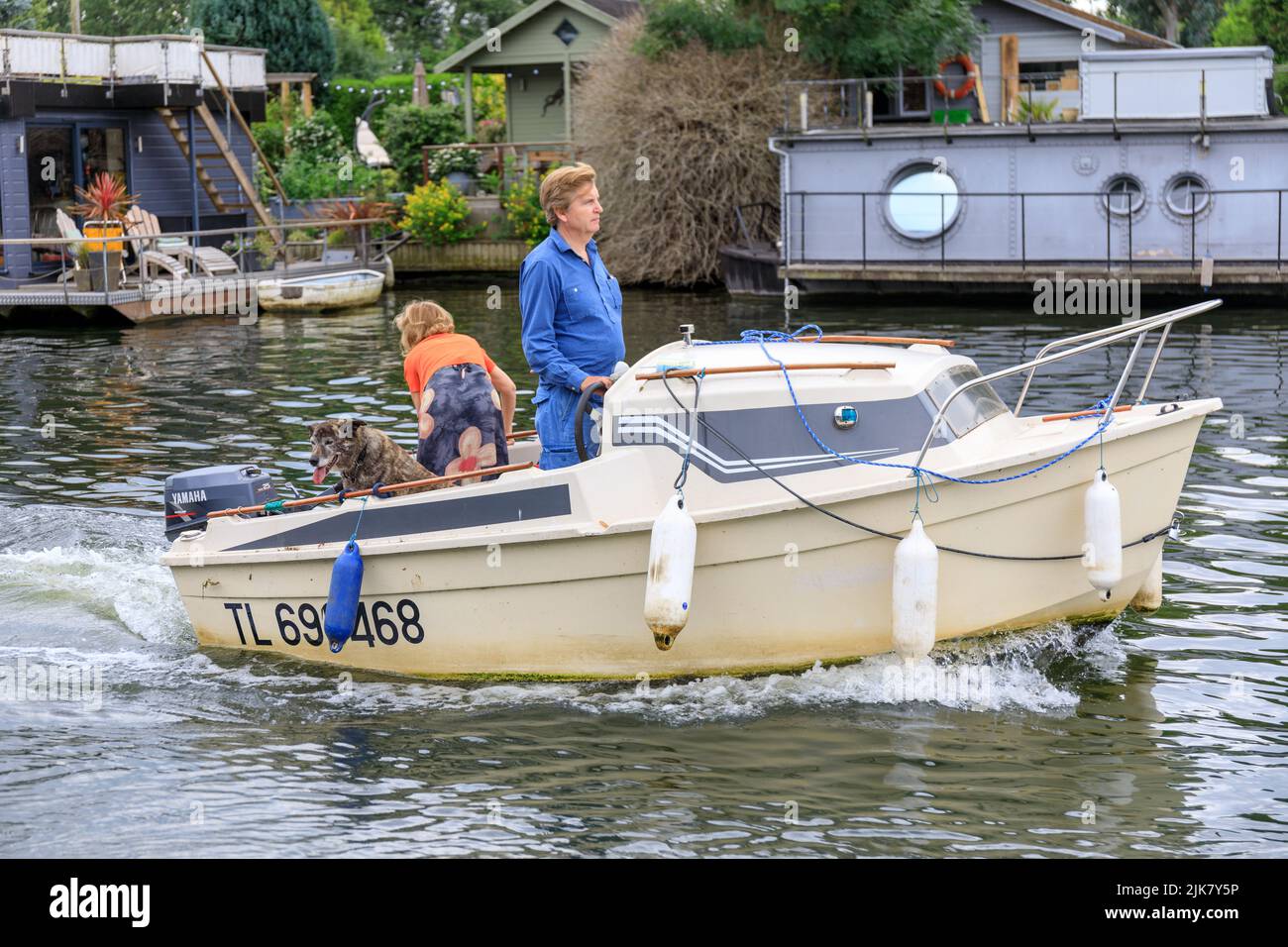 Small white cabin cruiser boat motors along the river Thames with a man steering the boat and a woman and a dog in the back. Houseboats in backgroung Stock Photo