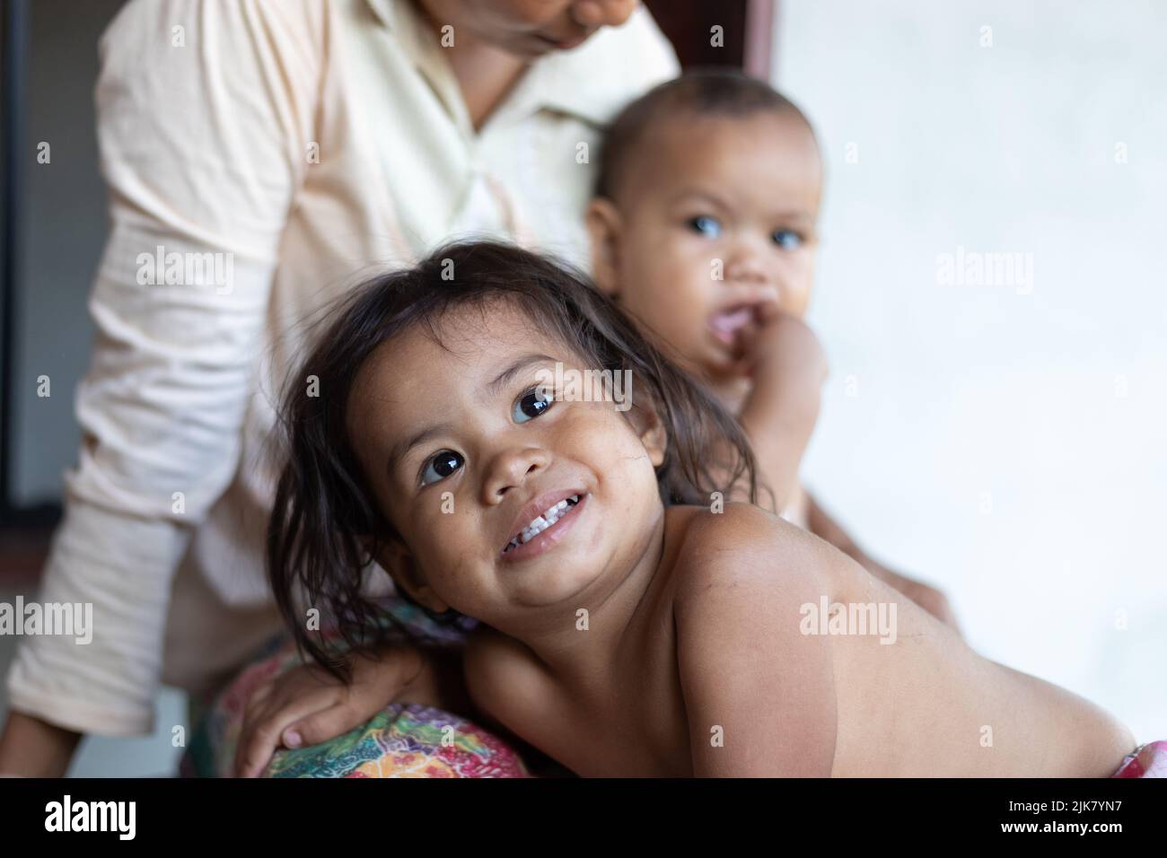 A young Thai girl smiles as she leans on her mothers lap with her baby sister looking on from behind Stock Photo