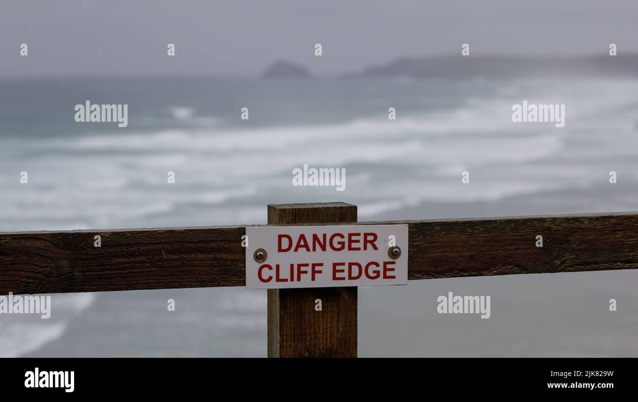 Danger cliff edge sign above a beach with waves and white water and distant headland, on a windy, wet day as a summer storm passes in Cornwall Stock Photo