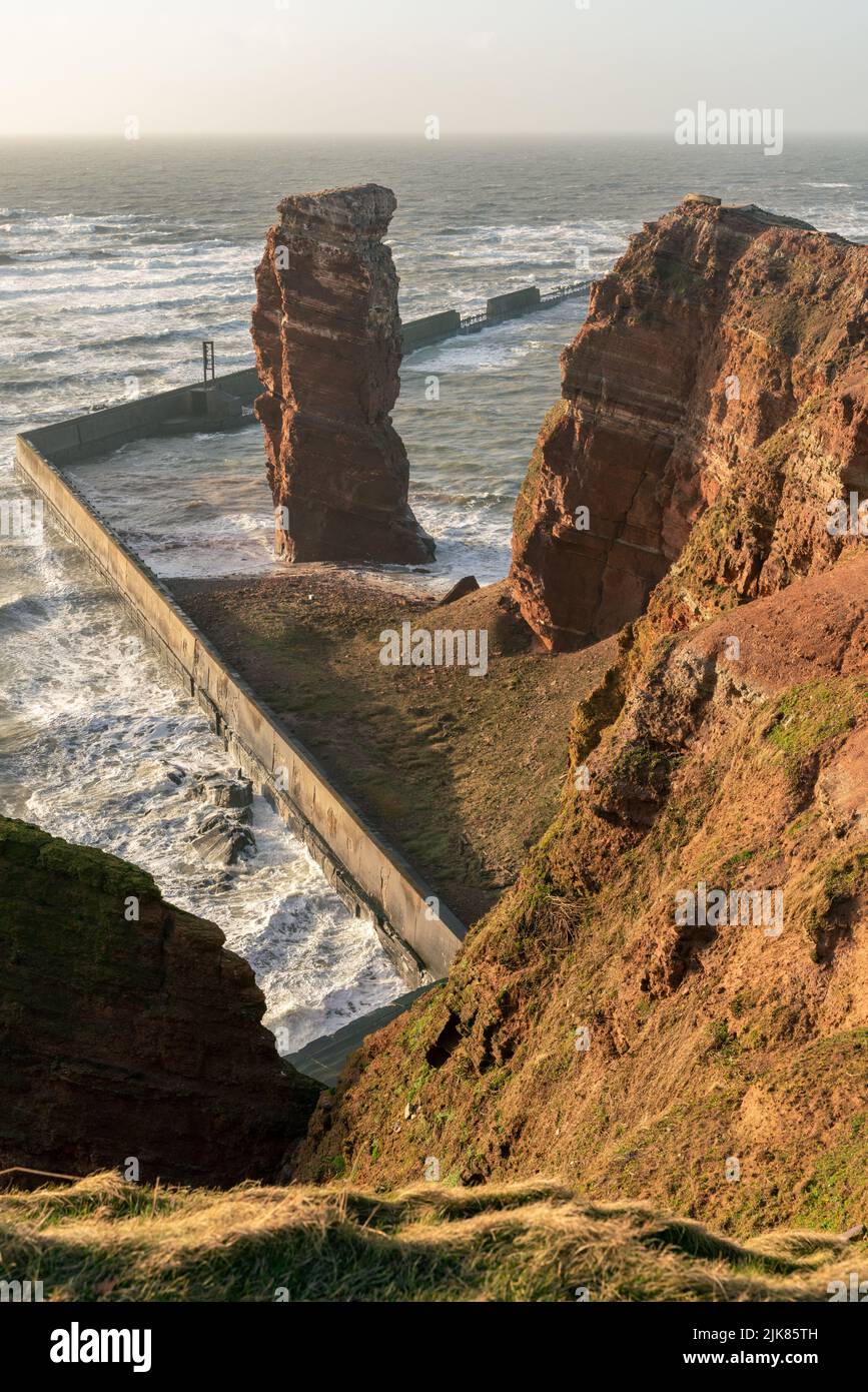 Lange Anna sea stack and tall red dramatic cliffs of Heligoland island with rough sea. Sunny windy evening in golden hour. Helgoland island in the Stock Photo