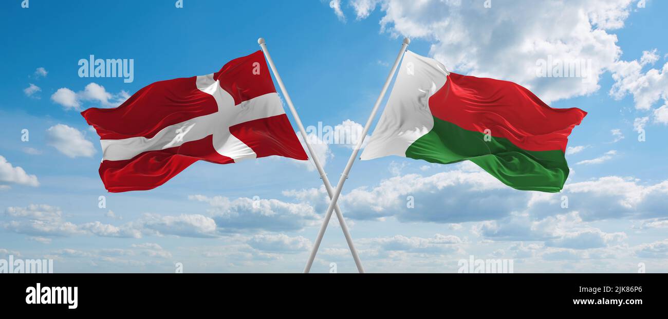 two crossed flags Madagascar and Denmark waving in wind at cloudy sky ...