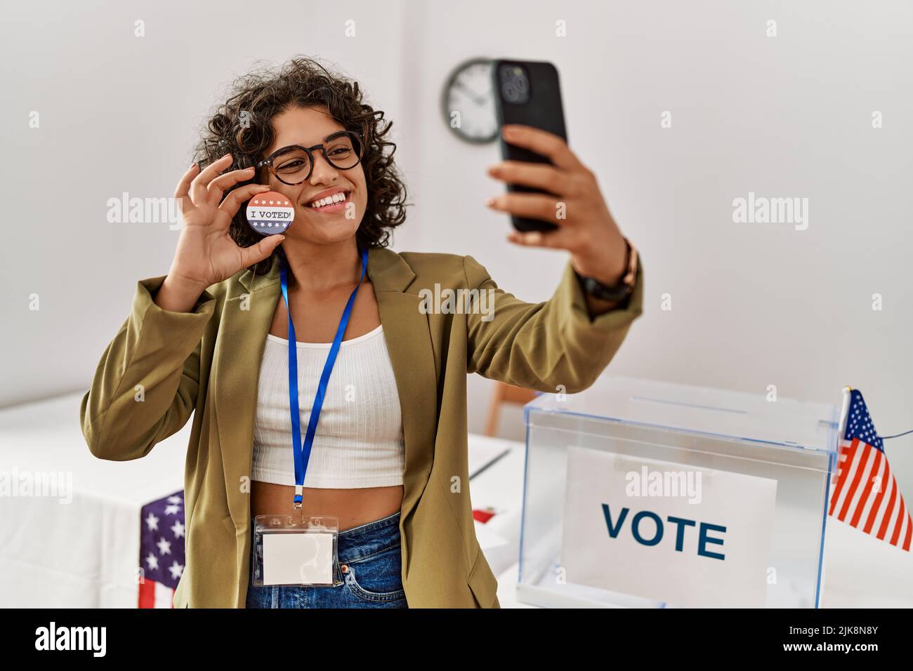 Young hispanic woman holding i voted badge make selfie by the smartphone at electoral college Stock Photo
