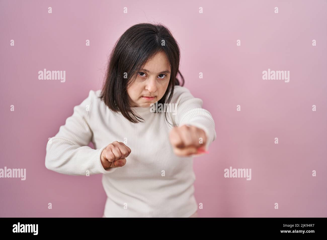 Woman with down syndrome standing over pink background punching fist to