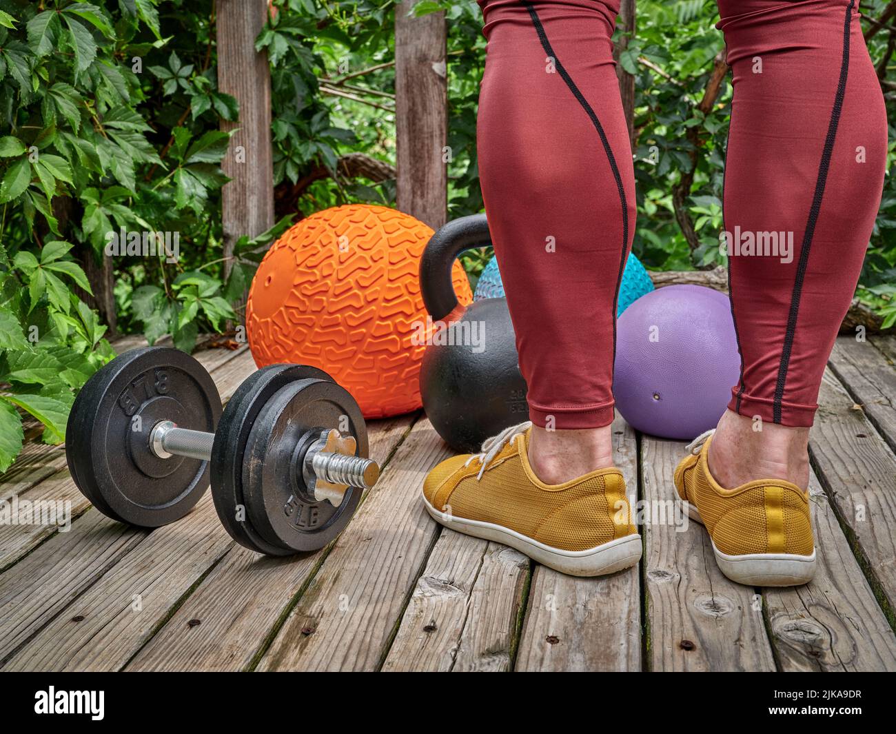 male in compression pants is exercising with heavy slam balls, dumbbell and kettlebell on a backyard deck, functional fitness and backyard gym concept Stock Photo
