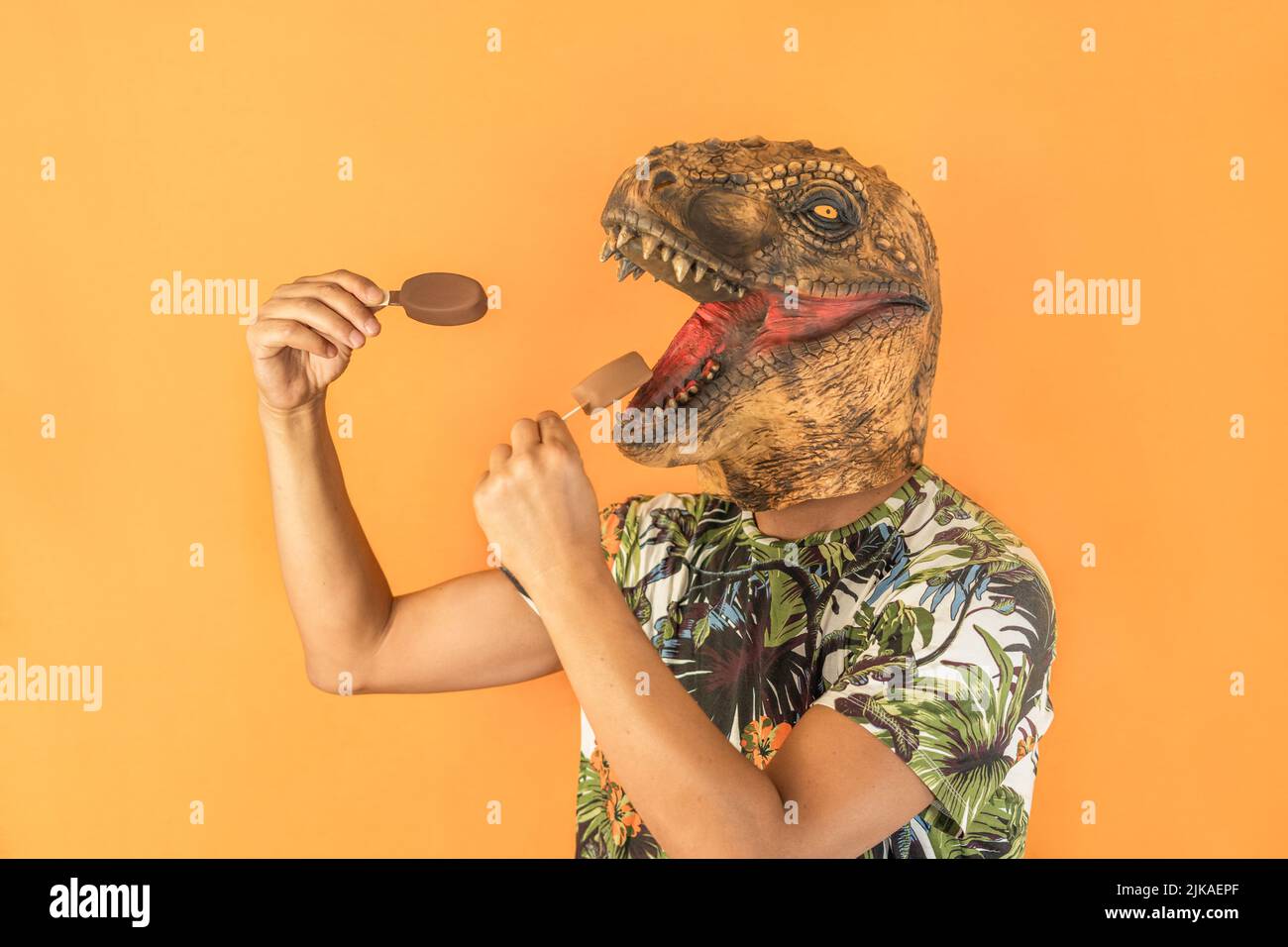 Man in dinosaur animal head mask eating two chocolate ice creams isolated on yellow background with copy space Stock Photo