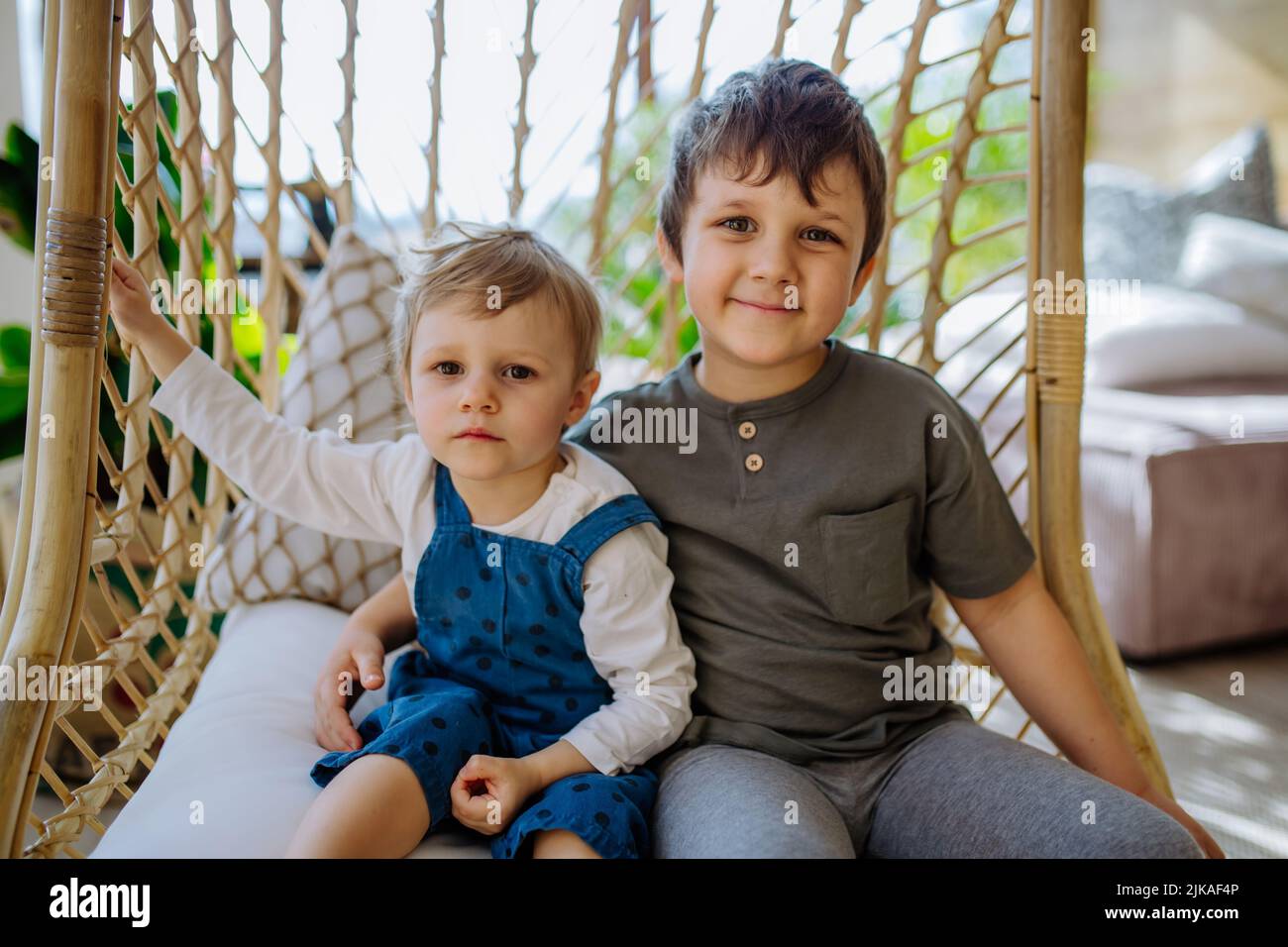 Little siblings playing, having fun and swinging on hanging chair in conservatory at home. Stock Photo