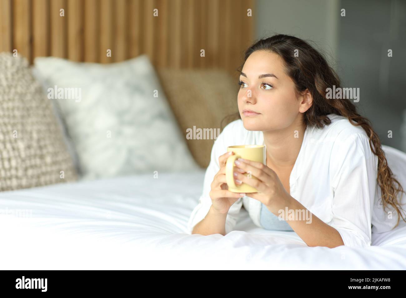 Distracted woman looking away drinking coffee lying on a bed in the bedroom Stock Photo