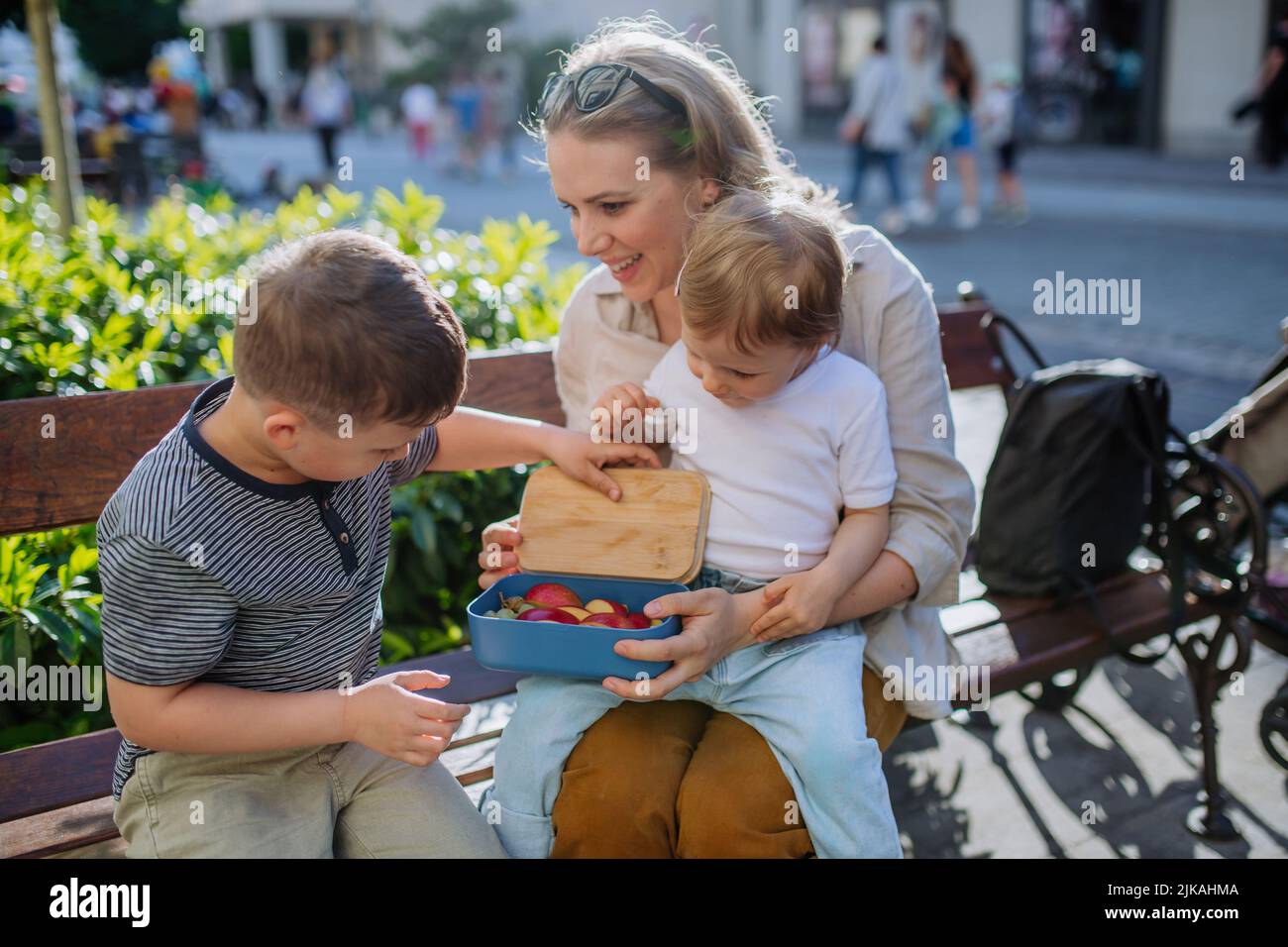 Young mother with little kids sitting on bench in city in summer, eating fruit snack and drinking water. Stock Photo