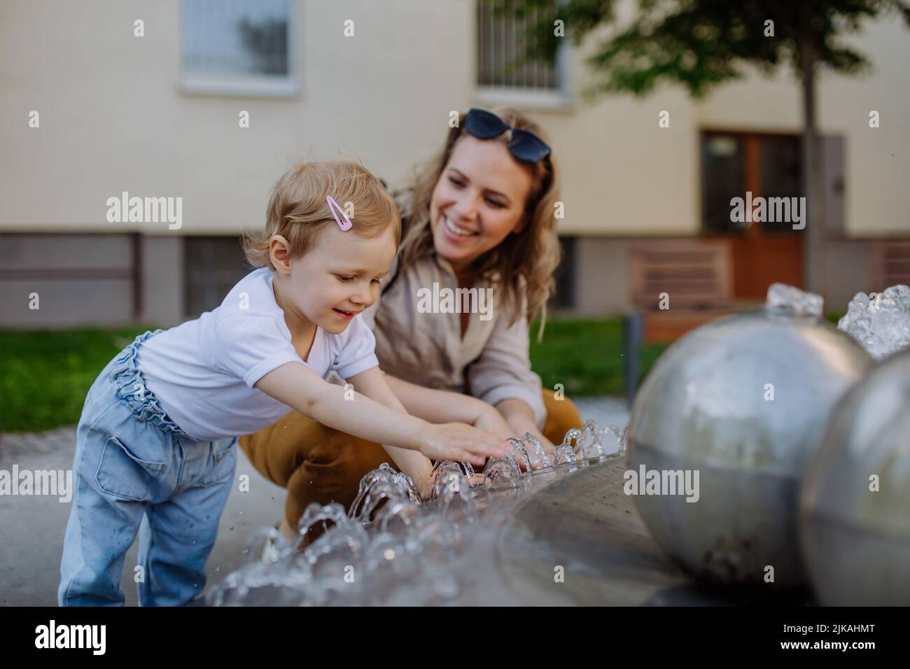 Young mother playing with her kid by fountain in city in summer. Stock Photo