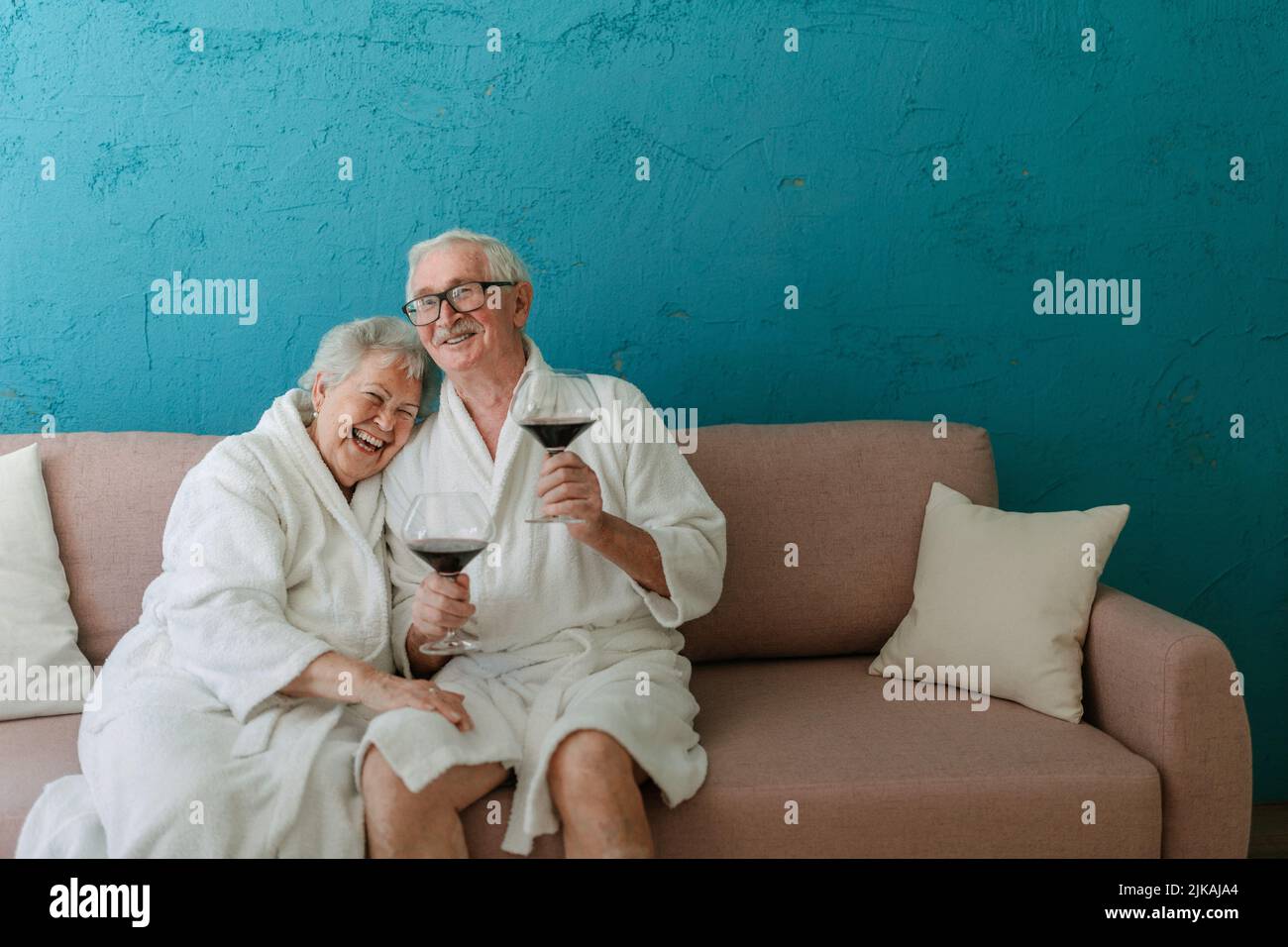 Happy senior couple sitting together in bathrobe on sofa with glass of wine, having nice time at home. Stock Photo