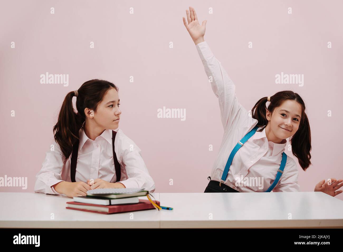 Enthusiastic perky schoolgirl raising a hand, her twin eyeing her with suspition Stock Photo