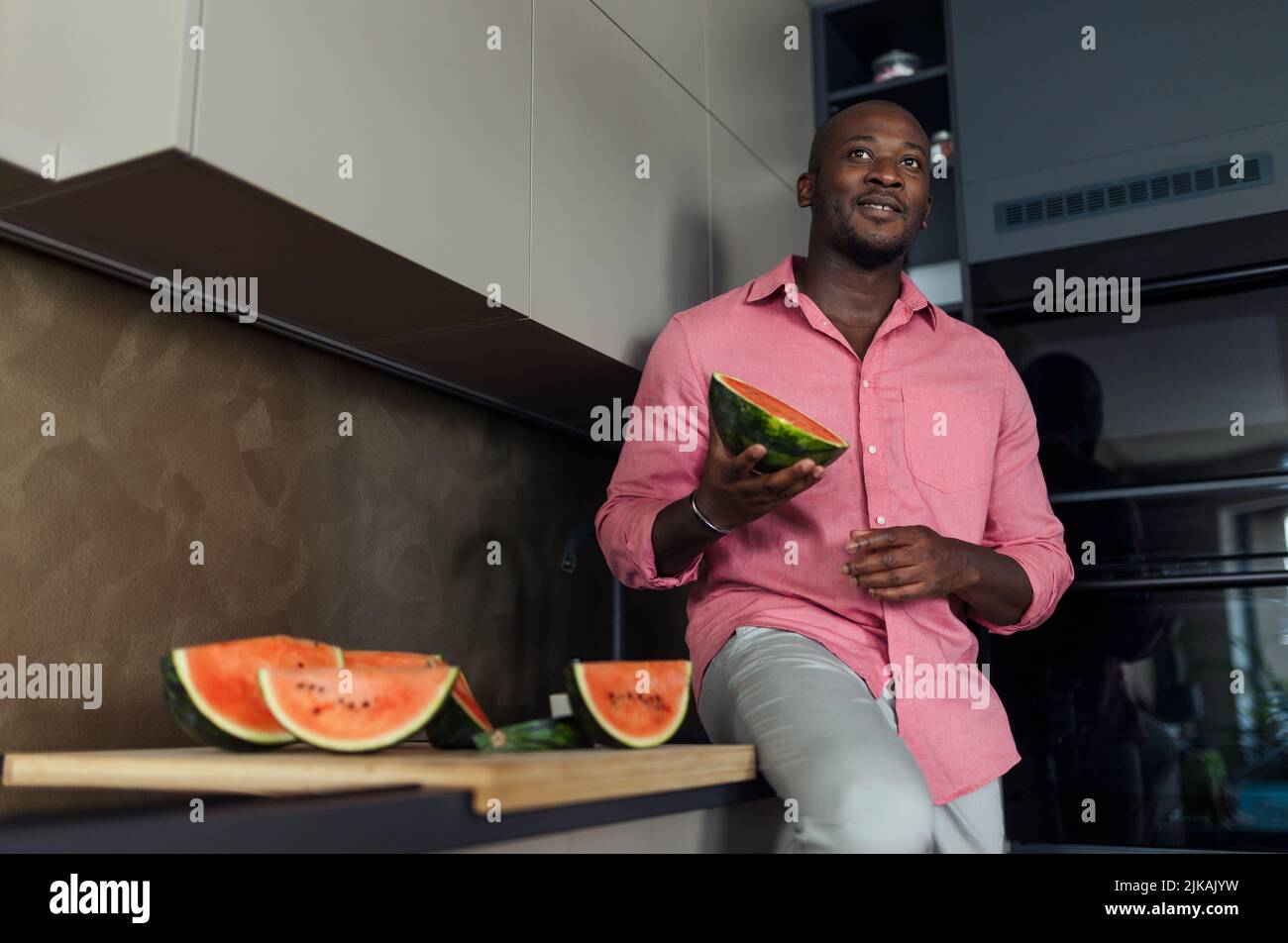 Multiracial man eating watermelon in his kitchen during hot sunny days. Stock Photo