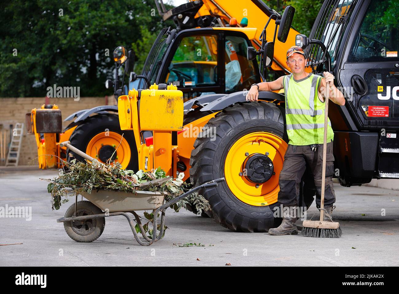 A yard manager posing with a wheelbarrow and yard broom next to a JCB Telehandler at a plant hire company Stock Photo