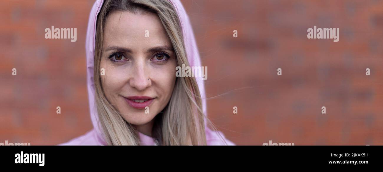 Wide photography of young blond woman with hood on head, standing in front of brick wall. Stock Photo