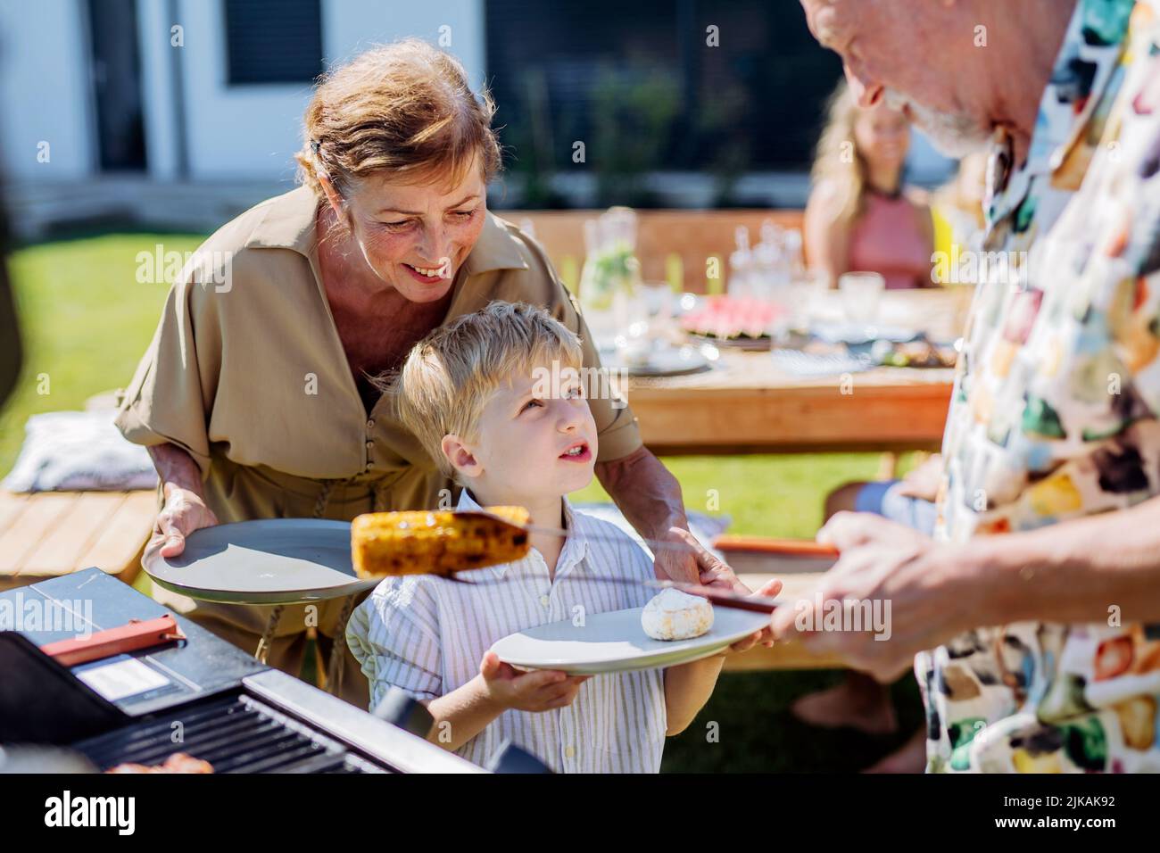 Grandparents giving grilled cheese and corn their grandson at garden grill party. Stock Photo