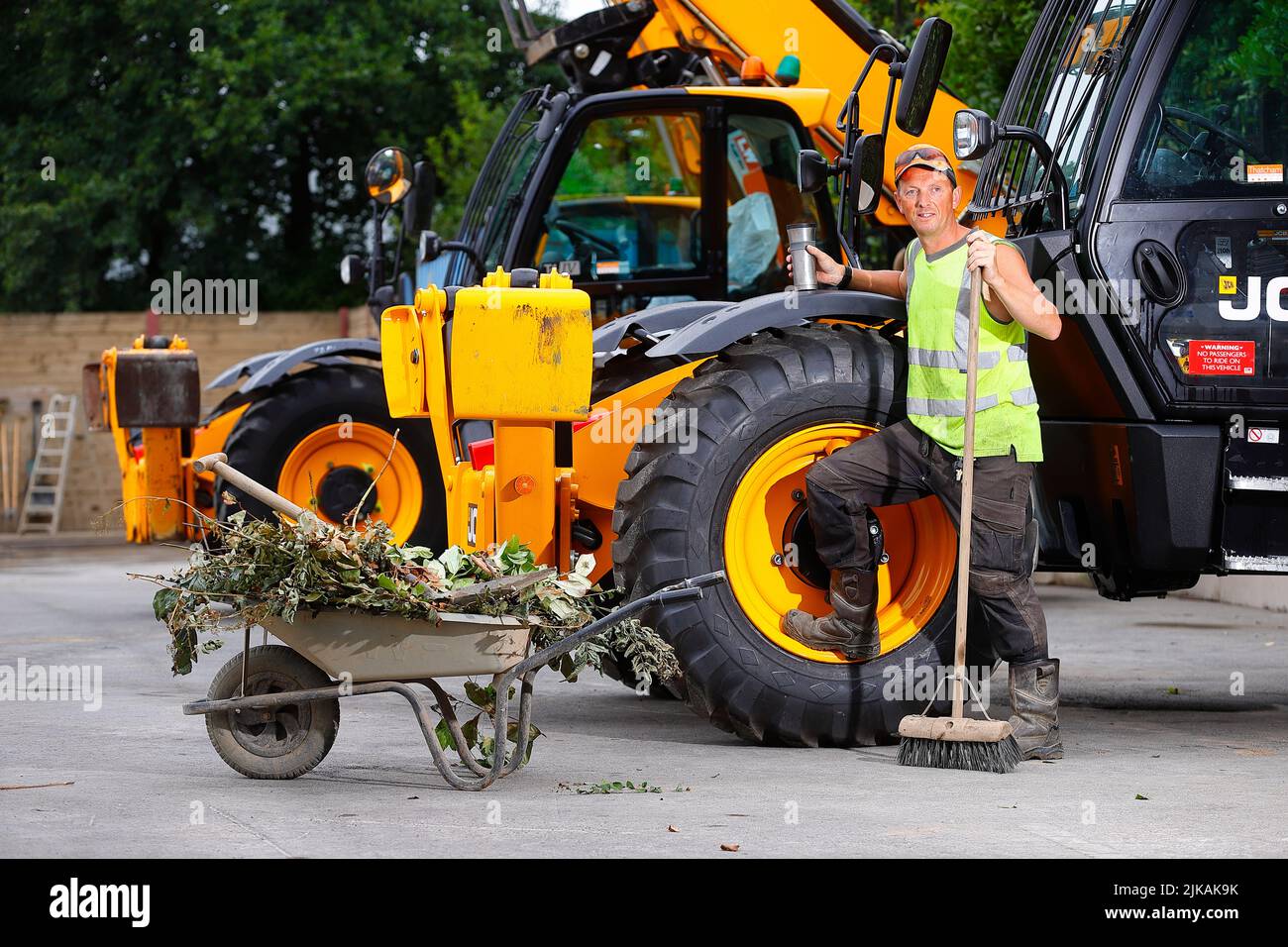 A yard manager posing with a wheelbarrow and yard broom next to a JCB