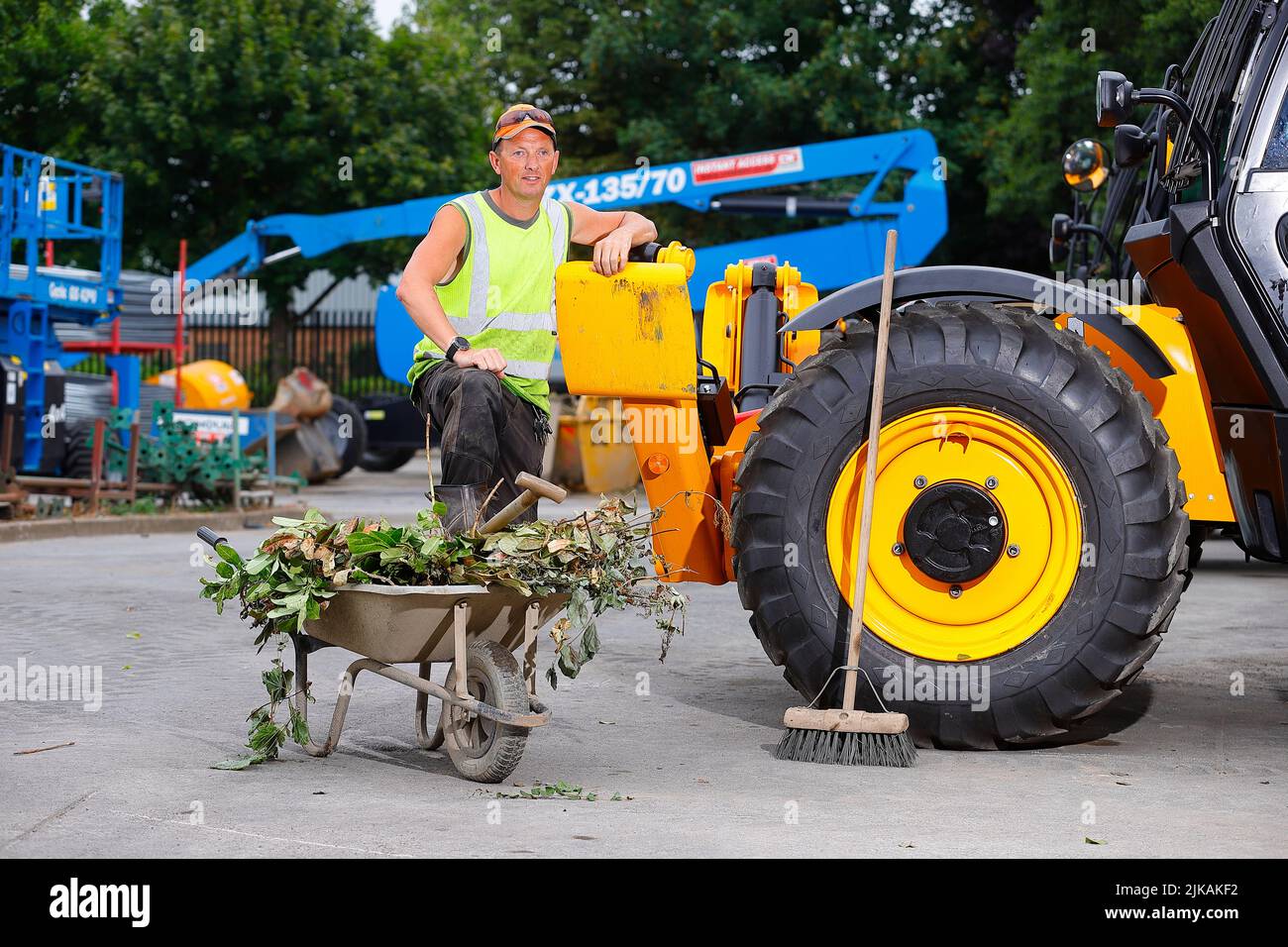 A yard manager posing with a wheelbarrow and yard broom next to a JCB Telehandler at a plant hire company Stock Photo