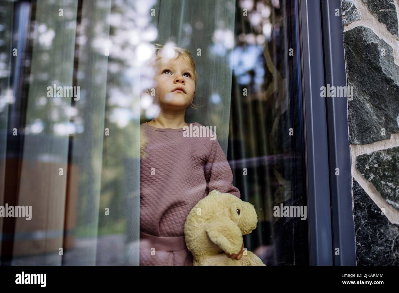 Little child standing alone with teddy bear behind the window, photo trough glass. Stock Photo