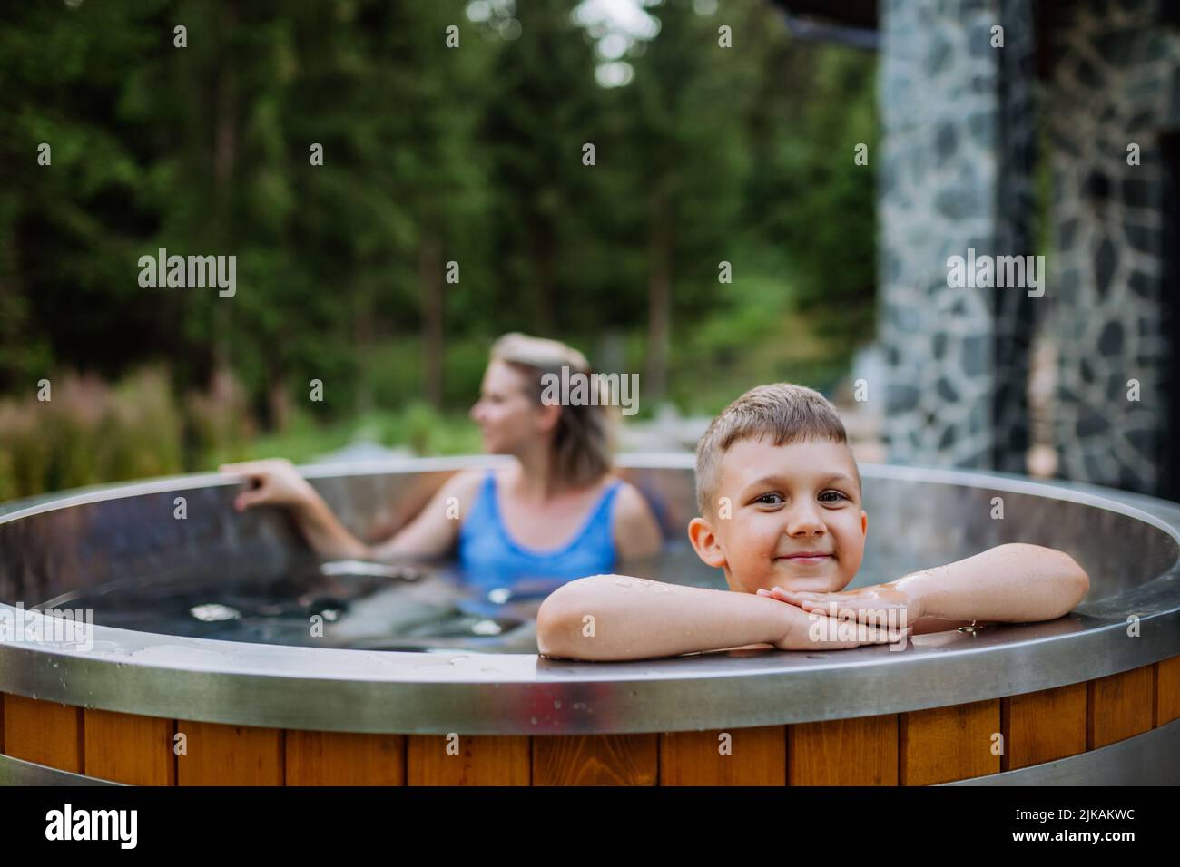 Mother with her little son enjoying bathing in wooden barrel hot tub in the terrace of the cottage. Wooden bathtub with a fireplace to burn wood and Stock Photo