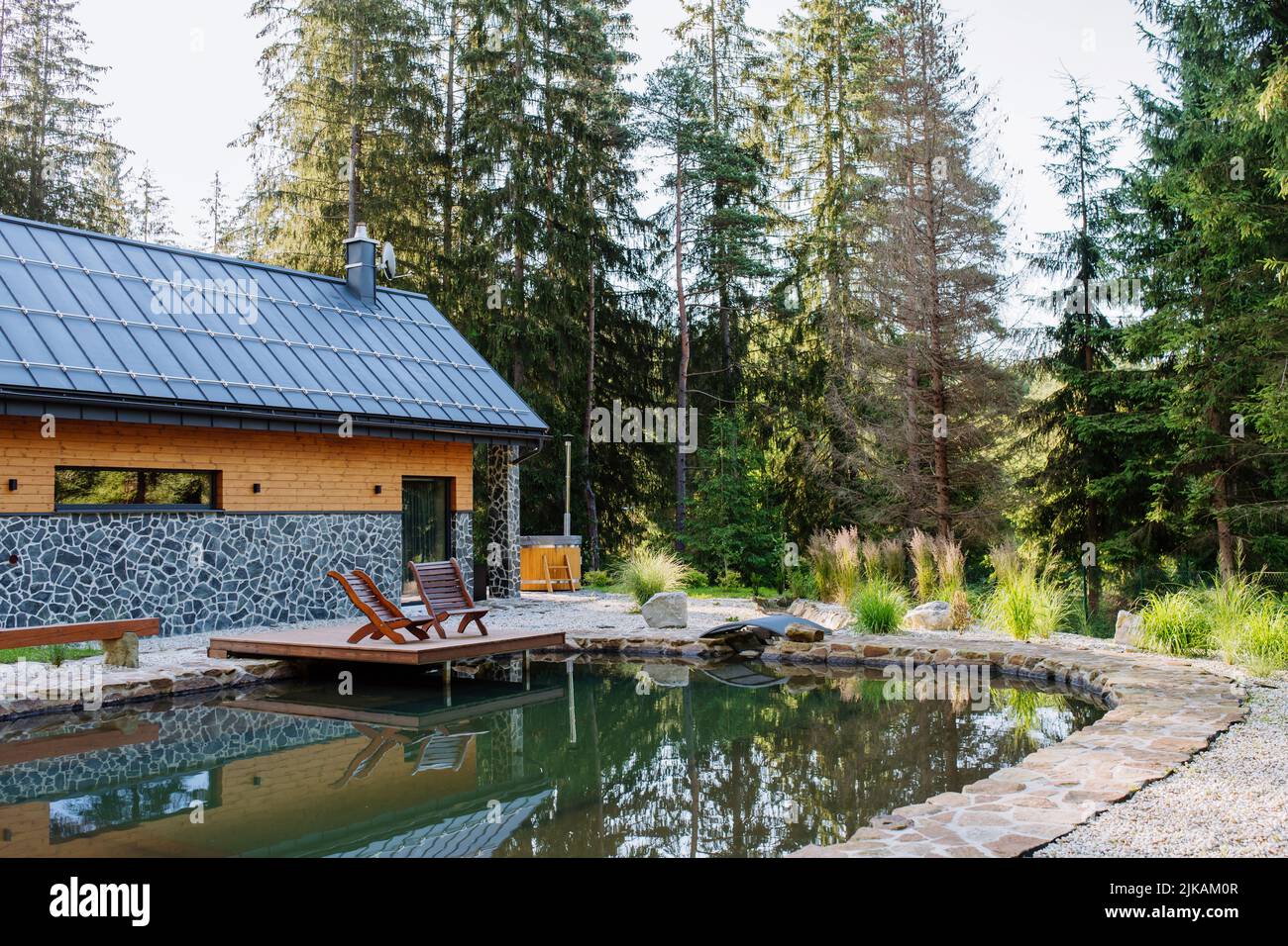 View of beautiful cottage in the middle of forest, surrounded by nature with natural pool and beach chairs. Stock Photo