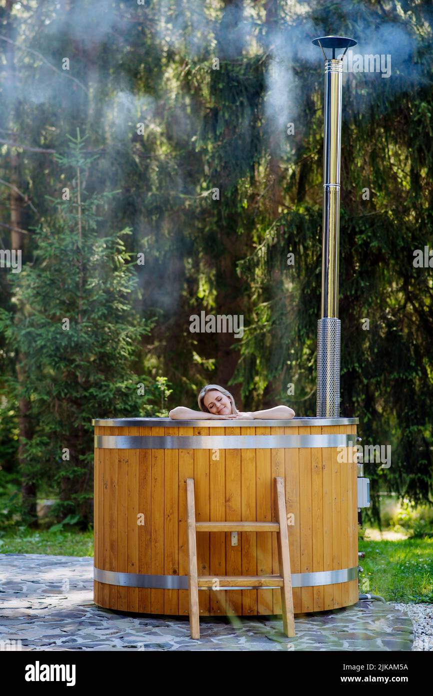 Young woman enjoying wooden bathtub with a fireplace to burn wood and heat water in backyard in mountains. Stock Photo