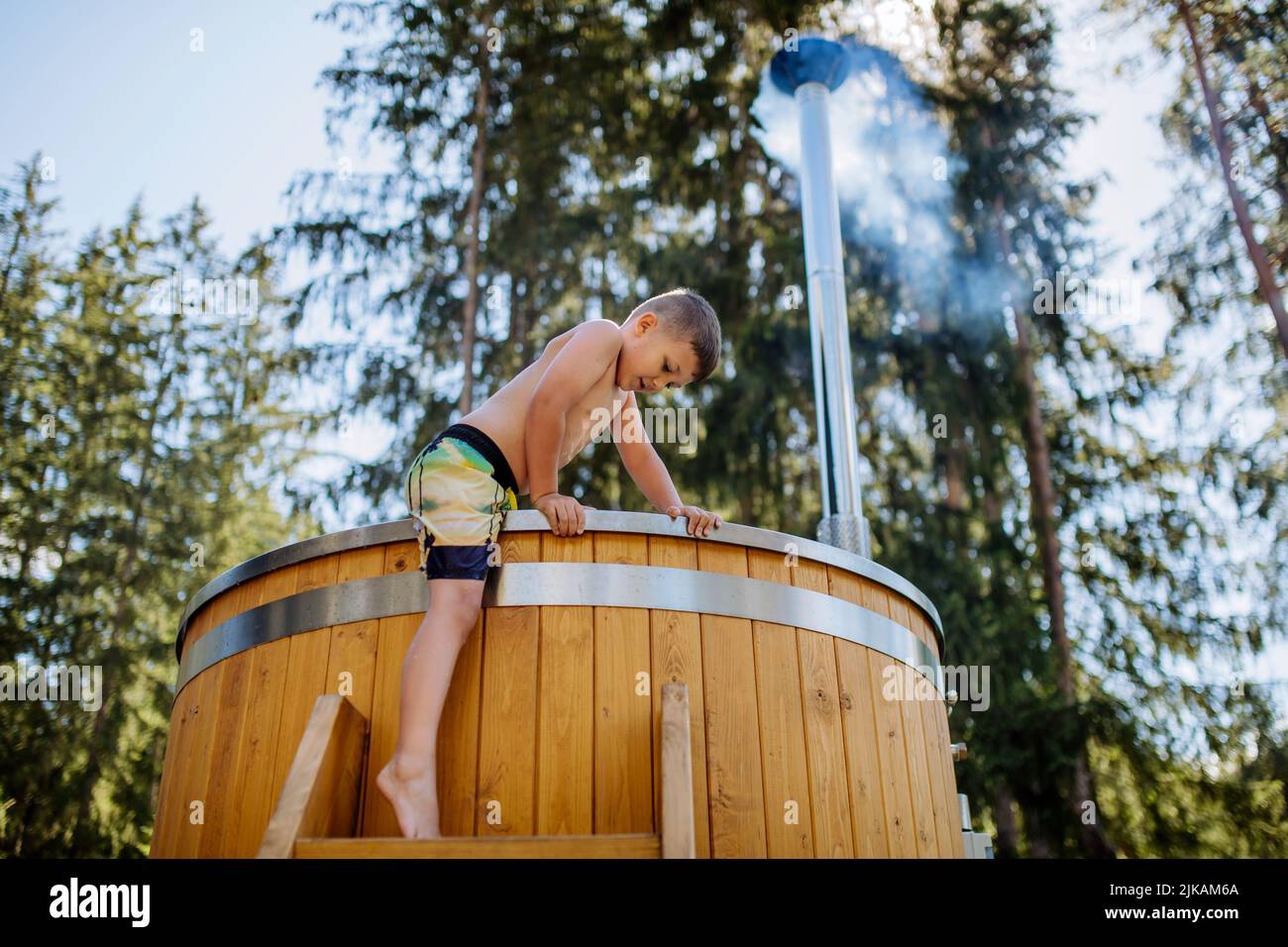 Little boy in swimsuit climbing in the outdoor wooden hot tub,surrounded by forest, during sunny summer day. Low angle view. Stock Photo