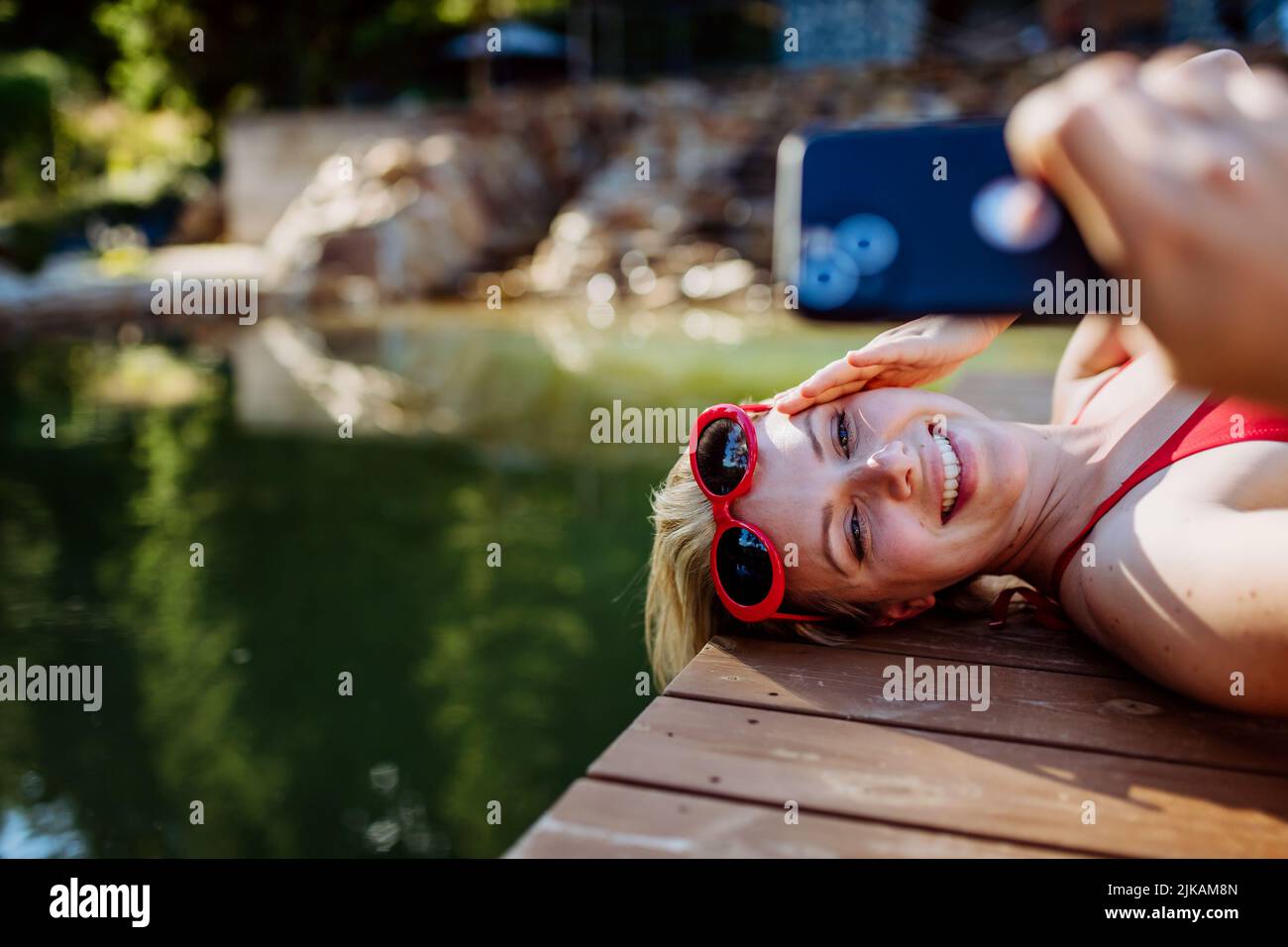 Rlaxed woman relaxing on the pier, lying and taking selfie near the natural lake in summer Stock Photo
